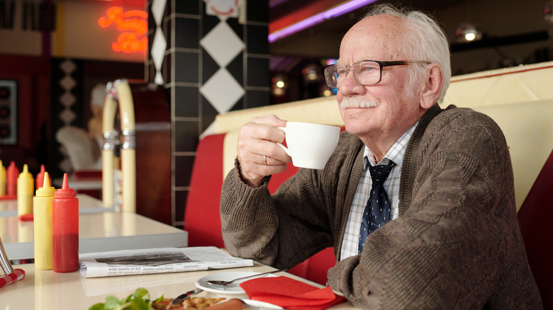 An elderly man sips a cup of coffee  in a retro diner with food in front of him.