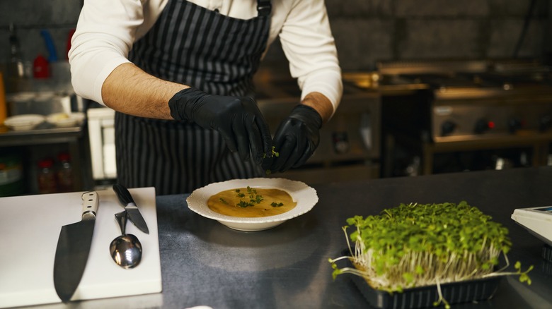 A chef in a black striped preparing a bowl of soup over a stainless steel countertop, with a cutting board and utensils on the side.