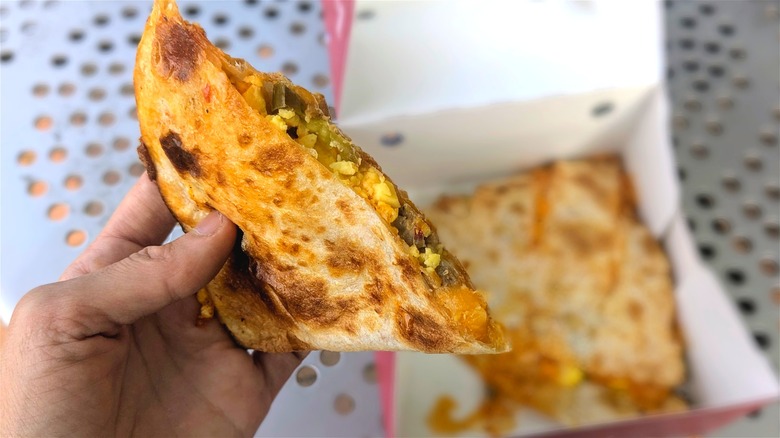 A hand holds a slice of a quesadilla while the rest of the quesadilla sits on a metal picnic table in the background