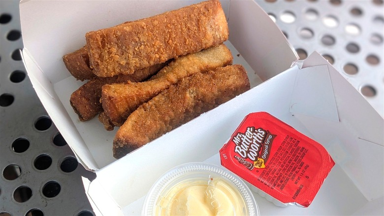 French toast sticks in a paper container with a syrup packet and cream cheese frosting cup