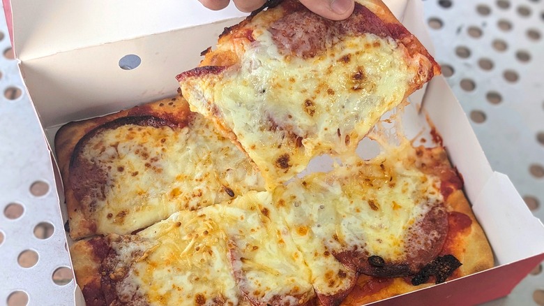 A hand lifting a slice of cheesy pizza flatbread from a paper box on a metal picnic table