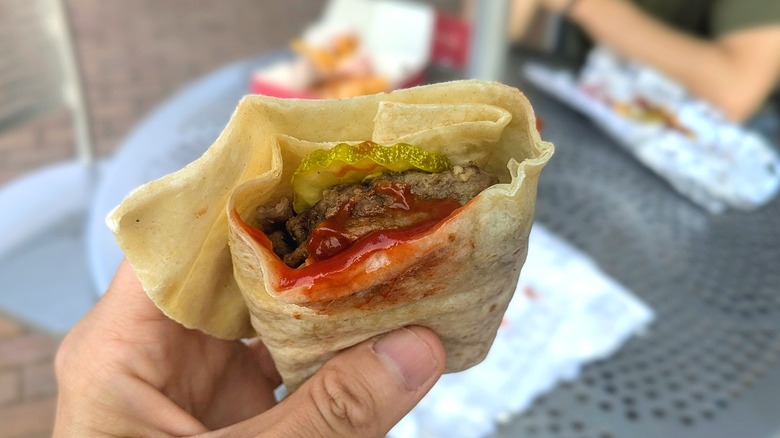 Close-up of a hand holding a wrap with pickles, ketchup, and a burger in front of a metal picnic table