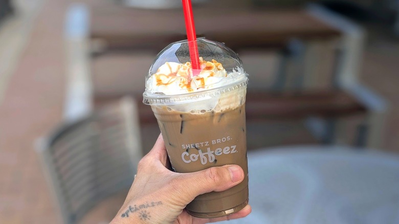 A hand holds an iced coffee with whipped cream in front of the picnic area of a Sheetz