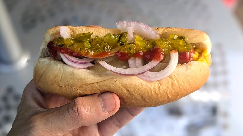 A hand holds a hot dog with onions and condiments