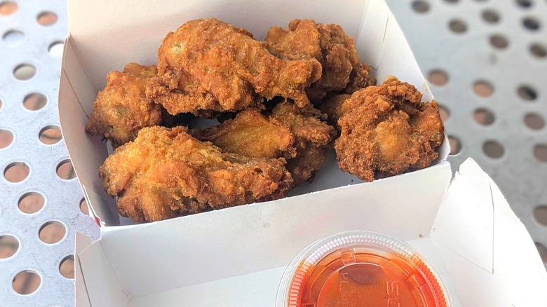 A dozen fried chicken wings in a paper container with a cup of hot sauce on a metal picnic table