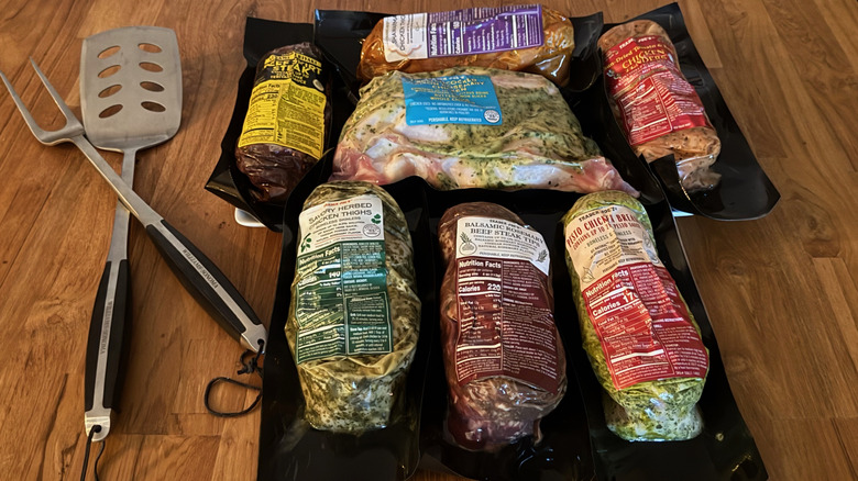 Seven varieties of Trader Joe's packaged marinated meats on a wood table next to a grilling spatula and fork.