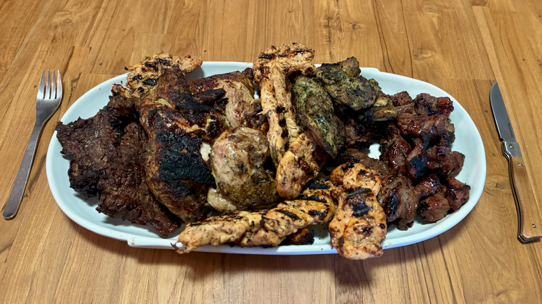 A large white platter filled with grilled meats on a wooden table between a fork and a steak knife.