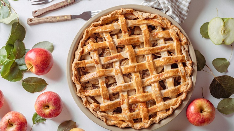 A lattice-topped apple pie surrounded by fresh apples