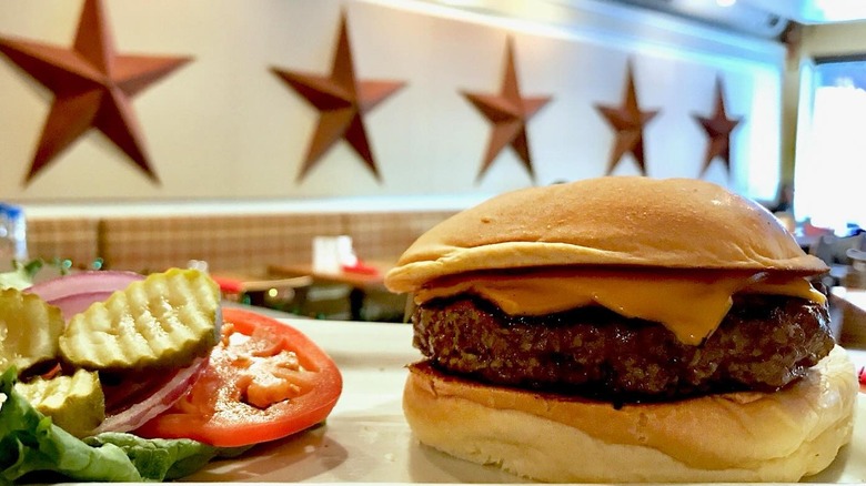 Five Star Burger on a tray next to a piece of lettuce, a tomato slice, a slice of red onion, and three pickle