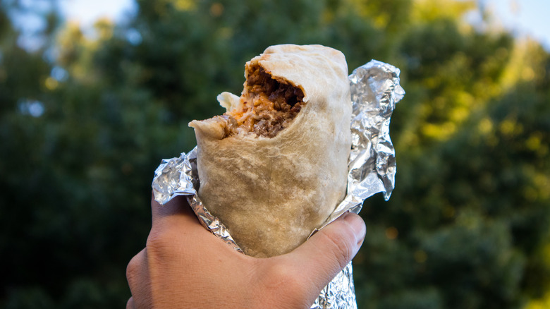 A person holds a burrito in foil with a bite taken out of it