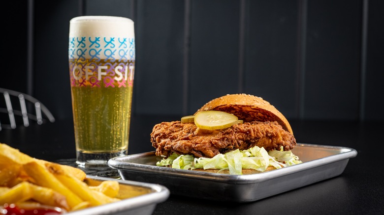 Offsite Miami Fried Chicken Sandwich with pickles and lettuce on plate beside a glass of beer and fries
