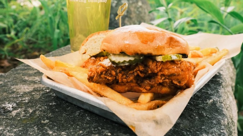 Rek-Lis Brewing Company Nashville Hot Chicken Sandwich, styled outdoors on a metal tray, a pint of beer and foliage in the background