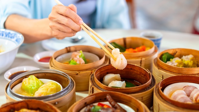 Hand with chopsticks taking shrimp from Chinese dim sum  steamer, surrounded by other dishes.