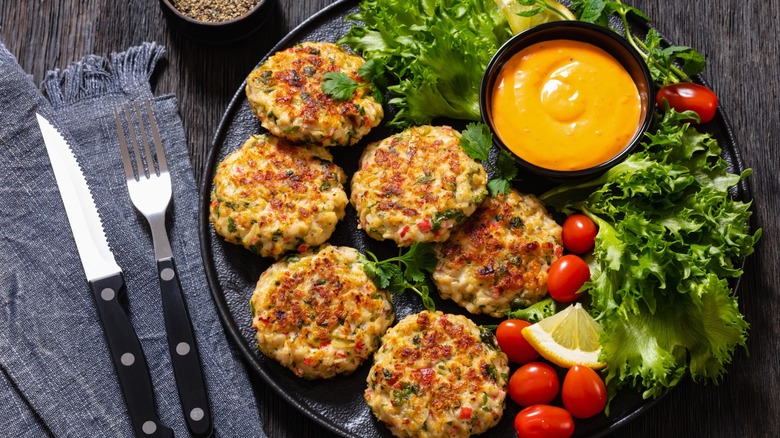 crab cakes with remoulade sauce, lemon, tomatoes, fresh lettuce on black plate on dark wood table, horizontal view from above
