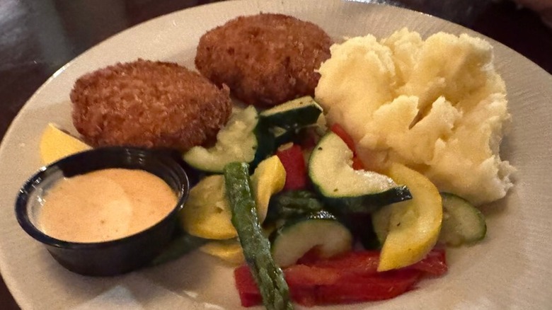 Crab cakes with sides of veggies, sauce, and mashed potatoes on a white plate at Waterman's Surfside Grill