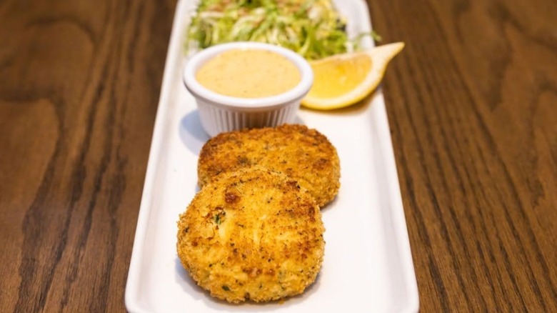 Crab cakes with sauce in white ramekin, lemon wedge on a rectangular white plate at Roosevelts