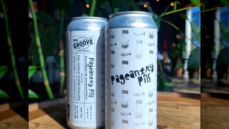Two tall white cans of Pageantry Pils beer on wooden surface