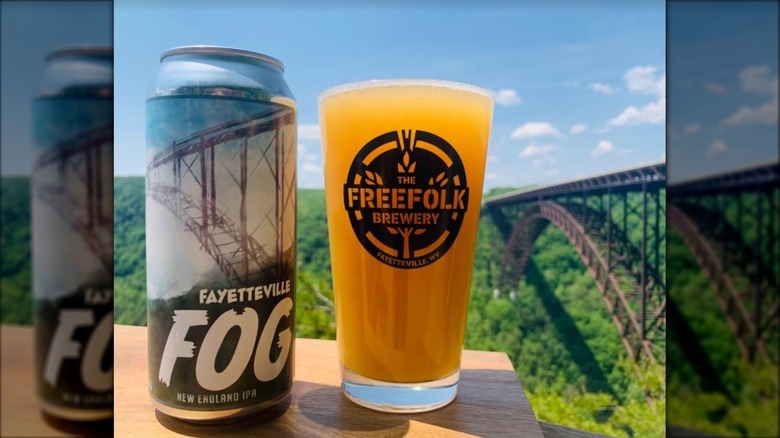Fayetteville tall beer can next to beer glass filled with orange beer, all outside near old metal bridge