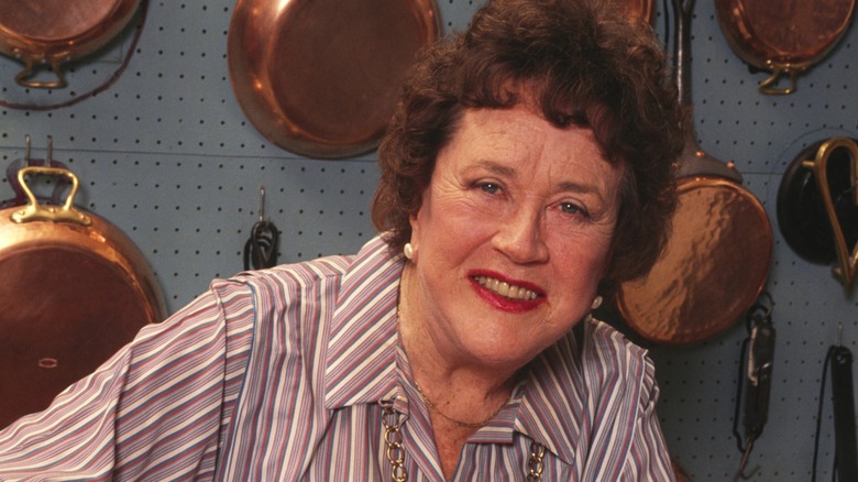 Julia Child smiles in her kitchen in front of a wall of copper pans