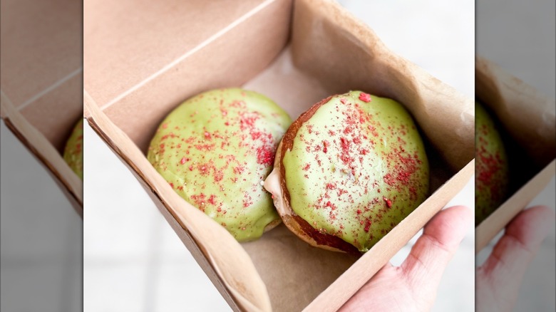 Hand holding box containing two iced donuts with green frosting and pink dusting