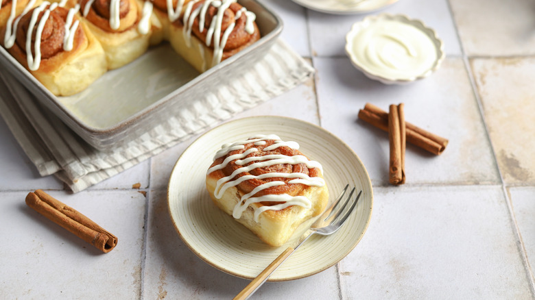 Baked cinnamon roll on a plate with frosting drizzled on top with forks, a pan, and frosting in background.