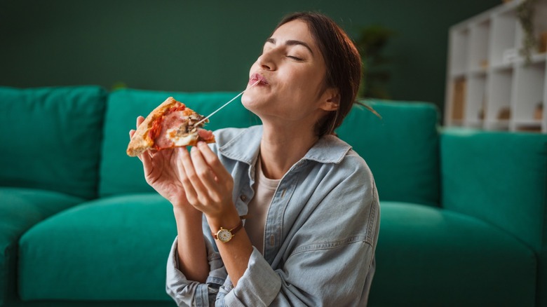 Young woman tasting a pizza slice in a living room, big cheese pull shown
