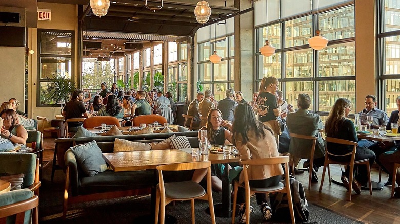 A bust restaurant scene with tables full of diners, glass windows and lamps hang to create a cozy ambiance