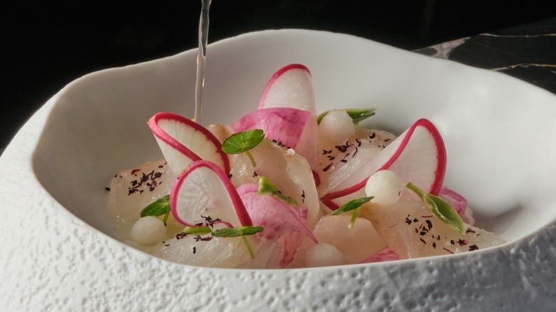 A close up of a white pitted bowl filled with slivers of radish and crudo and olive oil being poured over