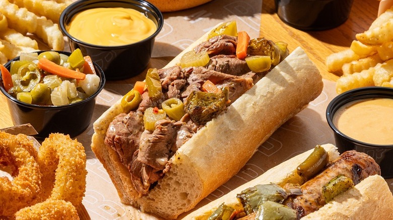 A picture of a buona beef sandwich, surrounded by fries, onion rings, garnishes and a sausage on a bun.