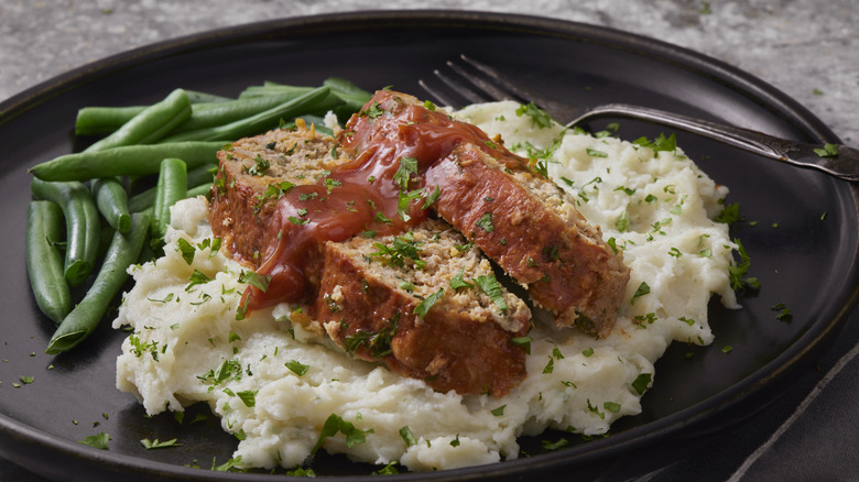 Two slices of meatloaf with sauce sitting on a bed of mashed potatoes on a black plate.