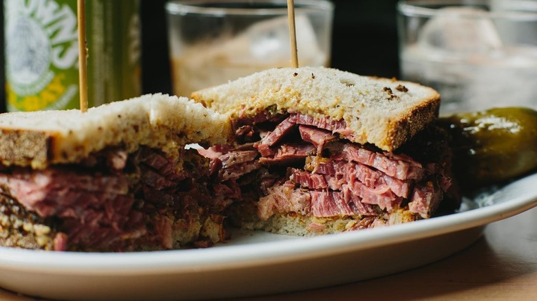 Pastrami sandwich split down the middle on white plate with pickle on side