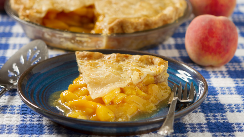 Slice of peach pie with a fork on a glass plate, with a peach in the background