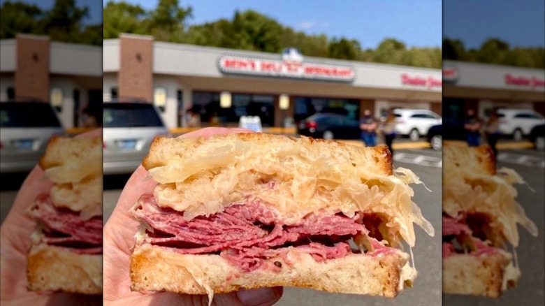 A hand holding a Reuben sandwich with restaurant in the background