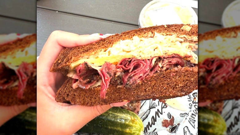 A hand holding a Reuben sandwich with chips and pickles on picnic table
