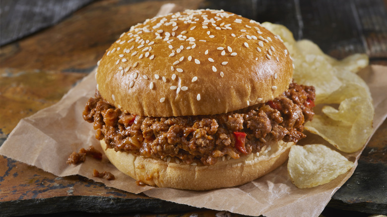 Sloppy joe on sesame seed bun on brown wax paper with side of potato chips