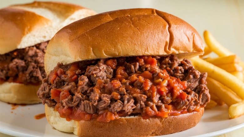 Two sloppy joes on white hamburger buns on white plate with side of french fries