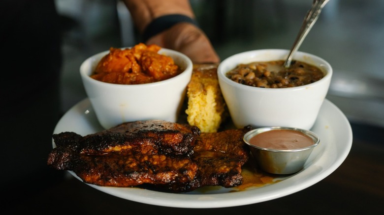 Hand holding plate with fried meat, dipping sauce and side of soup, cornbread, and saucy vegetables