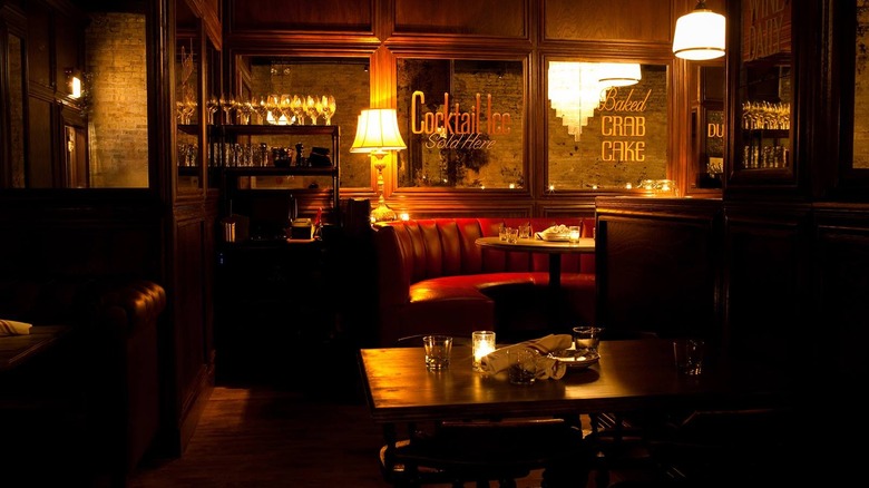 A dimly lit restaurant scene with red leater couch, lamps, tables with glasses on them, and wooden paneled walls