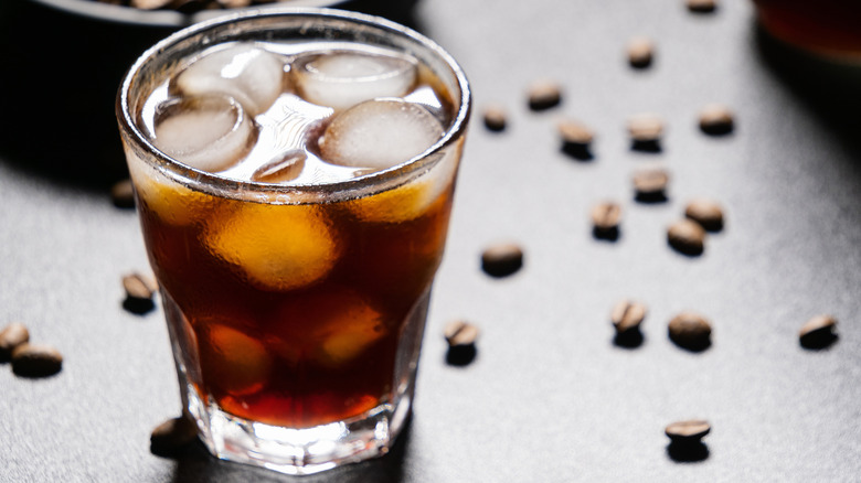 A glass of cold brew coffee with ice on a dark surface near coffee beans