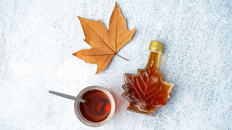 Maple syrup in glass bottle next to a maple leaf and a small bowl of syrup with a spoon in it.