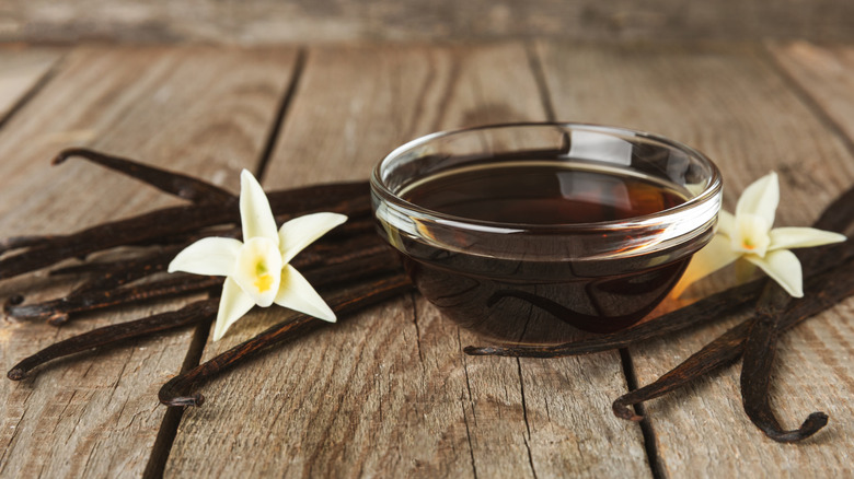 Small glass bowl of vanilla extract alongside vanilla bean pods and flowers