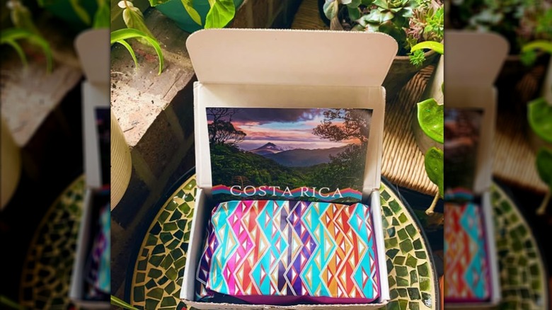 An open box on a green table displaying a postcard and a colorful bag