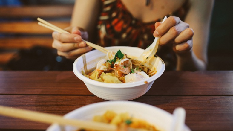 Close up image of diners eating two bowls of traditional Thai food, a pad Thai with shrimp and red curry noodles with chicken and vegetables. Served on a wooden table