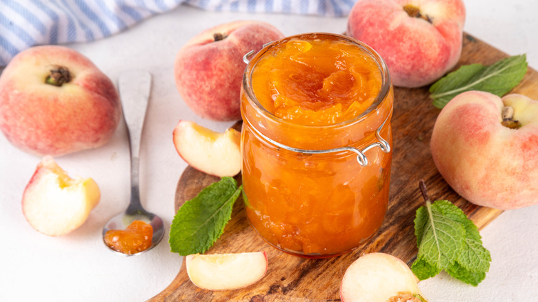 Jar of peach preserves on a wooden cutting board, surrounded by fresh peaches