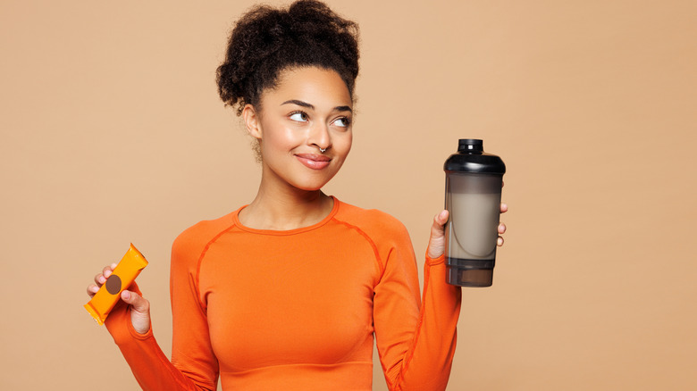Woman wearing holding a protein shake in one hand and a protein bar in the other, looking up to the side.