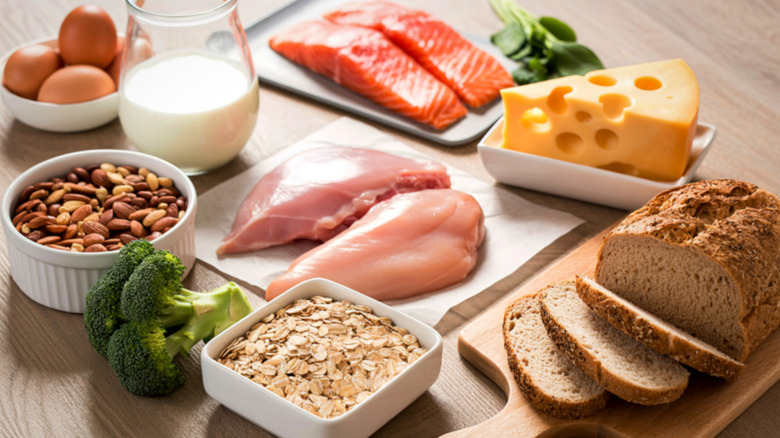 Sources of raw protein and carbohydrates: raw chicken breast, salmon, glass of milk, triangle of cheese, bowl of eggs, loaf of sliced whole grain bread, bowl of oats, bowl of mixed nuts and broccoli florets