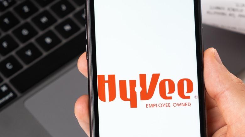 A hand holds a smartphone with the Hy-Vee app onscreen