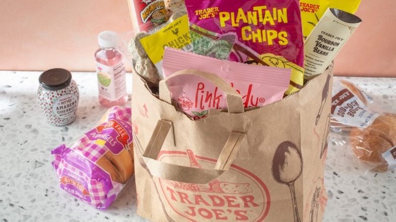 Paper Trader Joe's bag filled with groceries sits on a countertop in front of a light pink wall