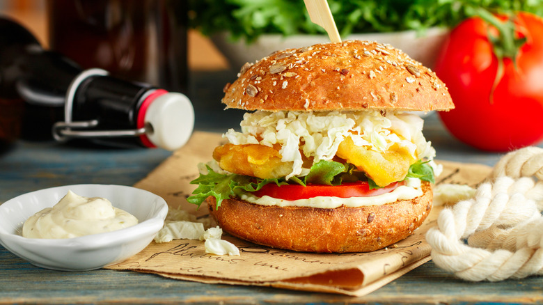 fried fish sandwich on paper with tartar sauce, lettuce, tomato, and cabbage