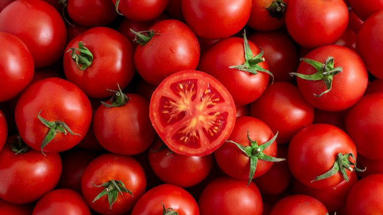 A sliced tomato sits in the center of a pile of whole, red tomatoes.